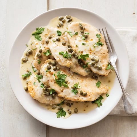 Golden pan-fried chicken breasts coated in flour, sizzling in a skillet with melted butter, ready to be smothered in a creamy lemon caper sauce for an easy dinner.  