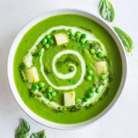 A bowl of vibrant green courgette, pea and pesto soup garnished with a dollop of crème fraîche and fresh basil leaves.